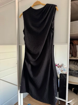 Svart asymmetrisk klänning H&M Divided - Svart klänning från H&M Divided med asymmetrisk design och draperad effekt. Klänningen är ärmlös och har en kort längd. Tillverkad i ett strukturerat syntetmaterial som ger en cool och modern look.