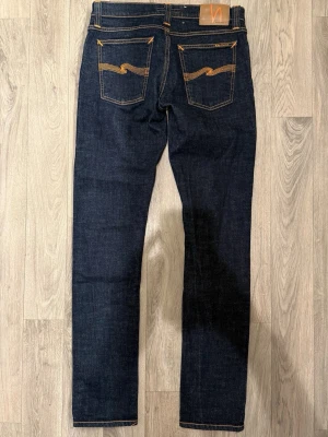 Nudie Jeans Long John Jeans Mörkblå Dry W29/L32 - Skinny Fit - Nudie Long John mörkblå dry jeans i stretchig denim med skinny fit passform som sitter tight och bekvämt och funkar till allt, storlek W29/L32, midja 37,5 cm, ytterbenslängd 102,5 cm, benöppning 15,5 cm, modellen är 185cm & väger 65kg, i fint skick.