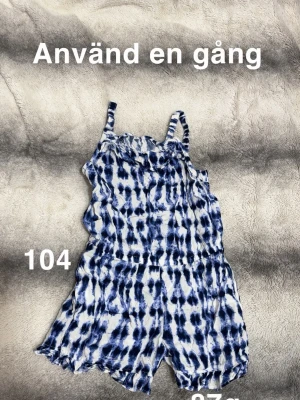 Blåvit tie-dye jumpsuit med volang - Snygg jumpsuit i blåvitt tie-dye-mönster med volangkant upptill och smala axelband. Modellen har resår i midjan och korta ben, perfekt för en avslappnad och trendig look. Mjuk och lätt i materialet.🌸 Använd en gång. Storlek 104