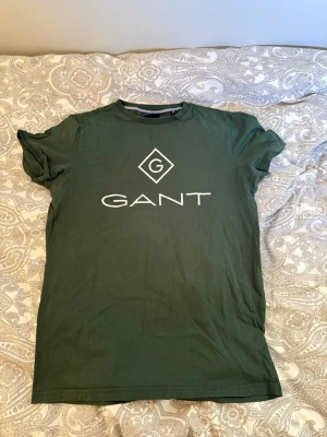 Grön GANT t-shirt med logga - Mörkgrön t-shirt från GANT med stor vit logga på bröstet. Klassisk rund hals och korta ärmar. Perfekt för en avslappnad och stilren look. Tillverkad i mjuk bomull som känns skön mot huden.