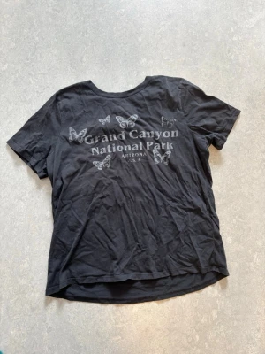 Mörkgrön Grand Canyon t-shirt - Säljer en mörkgrön t-shirt med tryck av fjärilar och texten 'Grand Canyon National Park Arizona USA' på bröstet. T-shirten har rund hals och korta ärmar. Materialet känns som mjuk bomull och passformen är avslappnad.