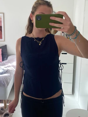 Mörkblå ärmlös topp med knyt i sidan - Snygg mörkblå ärmlös topp med rak halsringning och knytband i sidorna. Helt oanvänd med lapp kvar.