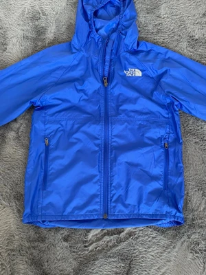 Blå vindjacka från The North Face - Snygg blå vindjacka från The North Face med huva och dragkedja framtill. Jackan har två fickor med dragkedja, elastiska muddar och reflekterande detalj på ryggen. Perfekt för blåsiga dagar och outdoor-aktiviteter.