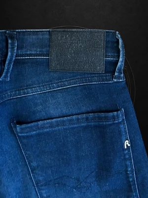  Replay Anbass - Snygga mörkblå slim fit jeans från Replay med klassisk femficksdesign och diskret logga på bakfickan. Jeansen har en modern passform och är tillverkade i stretchigt denimtyg för extra komfort. Perfekta för en trendig och avslappnad stil.
