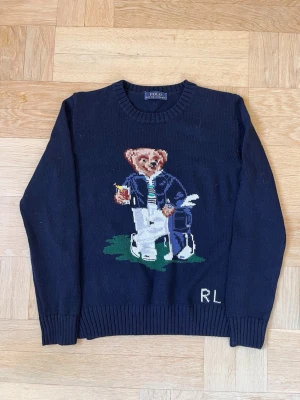 Mörkblå stickad tröja Polo Bear - Mörkblå stickad tröja från Polo Ralph Lauren med ikoniska Polo Bear-motivet framtill. Björnen har på sig en blå jacka, vit skjorta, randig slips och vita byxor, samt håller en drink. RL-broderi nere till höger. Rund hals och ribbade muddar.