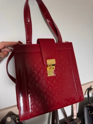 Röd  handväska lack - Snygg röd handväska  i glansigt lackläder med präglat mönster. Väskan har en fyrkantig form, dubbla handtag och guldfärgat lås framtill. Perfekt för dig som vill sticka ut med en exklusiv accessoar.