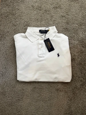 Vit pikétröja från Polo Ralph Lauren - Storlek S.Klassisk vit pikétröja från Polo Ralph Lauren med broderad logga på bröstet. Tröjan har krage, knäppning med tre knappar och är tillverkad i bomull. Perfekt för en stilren och tidlös look.