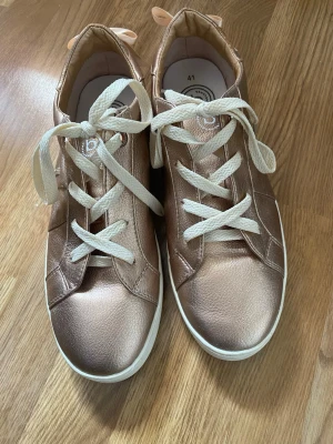 Roséguld,brons bugatti sneakers med beiga snören - Snygga Bugatti sneakers i roséguld, (brons )med beiga snören och vit sula. Skorna har en klassisk låg modell och är tillverkade i ett glansigt, läderliknande material. Perfekta för dig som vill sticka ut med en trendig och fräsch look.