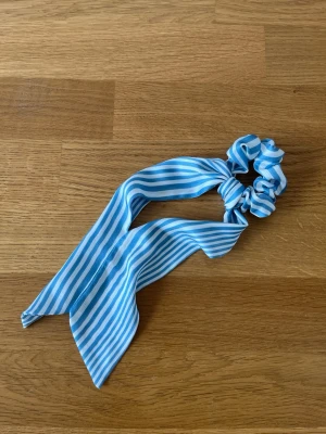 Blåvit randig scrunchie med rosett - Säljer en scrunchie i blått och vitt med smala ränder och en fastsydd rosett som ger en extra söt look. Tillverkad i ett mjukt tyg som känns skönt i håret. Perfekt för att piffa till din frisyr med en trendig accessoar.
