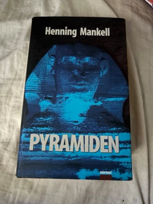 Pyramiden av Henning Mankell - En spännande kriminalroman av Henning Mankell med titeln 'Pyramiden'. Omslaget är svart med en blå Sfinx och tydlig titel i vitt. Boken är inbunden och ges ut av Ordfront. Perfekt för dig som gillar deckare och Wallander-serien.