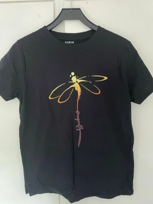 Svart t-shirt med trollslända  - Svart t-shirt från SHEIN med färgglad trollslända tryckt på bröstet. T-shirten har rund hals och korta ärmar. Materialet känns mjukt och passar perfekt till en avslappnad stil.