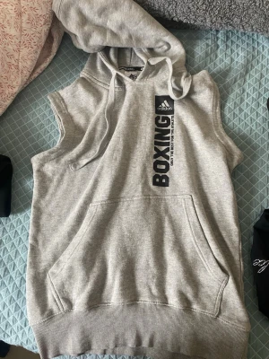 Grå ärmlös hoodie från Adidas - Säljer en grå ärmlös hoodie från Adidas med trycket 'BOXING'. Hoodien har en stor magficka och snören i huvan. Perfekt för träning och sportiga aktiviteter. Den är i väldigt bra skick 