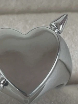 Silvrig hjärtring med pil - Cool ring i silverfärgad metall med ett hjärta som motiv och små pilar på toppen. Ringen har en öppen baksida vilket gör den justerbar i storlek. Perfekt accessoar för dig som gillar edgy och unika smycken. Rostfri 