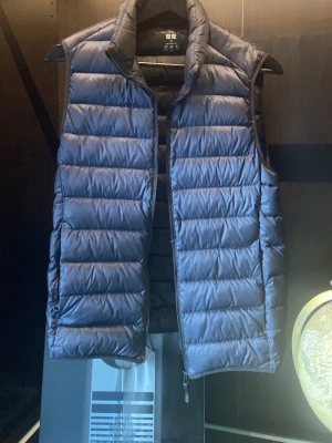 Marinblå dunväst från Uniqlo - Snygg marinblå dunväst från Uniqlo med hög krage och quiltad design. Västen har dragkedja framtill och är lätt vadderad för extra värme. Perfekt att slänga över en hoodie eller tröja för en cool lager-på-lager-look. I nyskick, kostar runt 1000 ny!