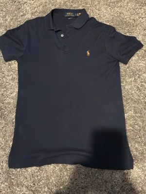 Marinblå pikétröja från Polo Ralph Lauren - Klassisk marinblå pikétröja från Polo Ralph Lauren med korta ärmar och krage. Tröjan har två knappar vid halsen och den ikoniska broderade loggan i gult på bröstet. Perfekt för en stilren och avslappnad look.
