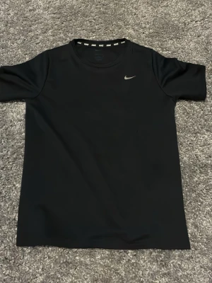 Svart Nike t-shirt i funktionsmaterial - Säljer en svart t-shirt från Nike med klassisk logga på bröstet. T-shirten är tillverkad i ett lätt och bekvämt funktionsmaterial, perfekt för träning eller en sportig look. Rund halsringning och korta ärmar ger en stilren och sportig vibe.