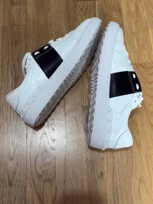 Valentino vita sneakers - Säljer ett par äkta Valentino Garavani Open sneakers i vitt läder med svart band. Storlek 45 men kan passa mindre. Skorna är i nytt skick, kommer med dustbag/kartong/kvitto. Nypris ca 6490kr