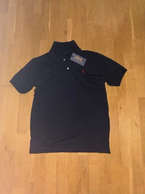 Mörkblå pikétröja från Polo Ralph Lauren - Klassisk mörkblå pikétröja från Polo Ralph Lauren med röd broderad logga på bröstet. Tröjan har korta ärmar, krage och knäppning med två vita knappar. Perfekt för en stilren och avslappnad look.