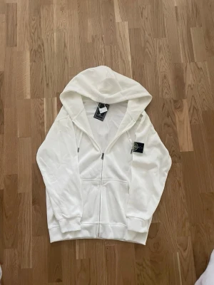 Vit hoodie från Stone Island - Snygg vit hoodie från Stone Island med dragkedja framtill och klassisk logopatch på vänster ärm. Hoodien har huva, ribbade muddar och känguruficka. Perfekt för en clean och stilren look. Materialet känns mjukt och bekvämt.