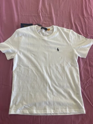Vit t-shirt från Ralph Lauren - Klassisk vit t-shirt från Ralph Lauren med den ikoniska broderade loggan på bröstet. T-shirten har rund halsringning och korta ärmar. Tillverkad i mjuk bomull för en skön och avslappnad känsla.