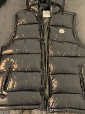 Svart dunväst  - Snygg svart dunväst från med glansig finish och klassisk logga på bröstet. Västen har dragkedja framtill, två fickor med dragkedja och en avtagbar huva. Perfekt för lager-på-lager och streetstyle. Nfc funkar och priset kan diskuteras vid snabb affär