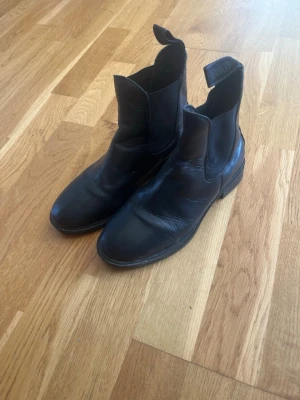Svarta boots  - Säljer ett par klassiska svarta  boots  i  läder. Skorna har elastiska paneler på sidorna och dragflikar både fram och bak för enkel påtagning. Tidlös design som funkar till det mesta och robust sula för bra grepp.