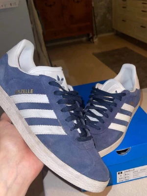 Adidas Gazelle blå sneakers - Blå Adidas Gazelle sneakers med klassiska vita ränder och vit häl. Skorna har en ovandel i mocka och vita detaljer på tungan och sulan. Guldtexten 'GAZELLE' på sidan ger en snygg touch. Perfekta för dig som gillar retrostil och ikoniska modeller.