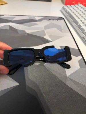 Off-White Solglasögon - Snygga solglasögon med rektangulära svarta bågar och coola blåtonade glas. Glasögonen har en modern, kantig design som sticker ut och passar perfekt till en trendig stil. Ramen är i plast och glasen har en mörkblå nyans.