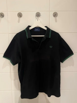 Svart pikétröja från Fred Perry - Klassisk svart pikétröja från Fred Perry med gröna detaljer på krage och ärmslut. Tröjan har korta ärmar, knappslå framtill och den ikoniska Fred Perry-loggan broderad på bröstet. Perfekt till en avslappnad och stilren look.