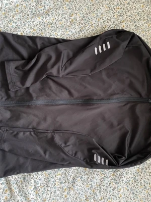 Svart sportjacka med reflexdetaljer - Säljer en svart sportjacka med hel dragkedja framtill och snygga reflexdetaljer på ärmarna. Jackan har en enkel, stilren design och är tillverkad i ett lätt och smidigt material som passar perfekt för löpning eller träning utomhus.