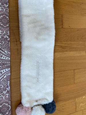 Vit fluffig halsduk med bollar - Supersöt vit halsduk i mjukt fuskpälsmaterial med tre fluffiga bollar i vitt, rosa och mörkblått i ena änden. Perfekt accessoar för att hålla dig varm och ge din outfit en gullig touch.