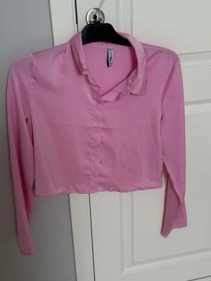 Rosa cropped blus med krage - Trendig rosa cropped blus med klassisk krage och knappar framtill. Blusen har långa ärmar och är tillverkad i ett tunt, lätt material som ger en avslappnad look. Perfekt att styla med höga jeans eller kjol.