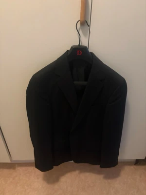 Svart kavaj från Dressmann Slim Fit - Stilren svart kavaj från Dressmann i modellen Slim Fit. Klassisk design med två knappar framtill, smala slag och innerfoder i svart. Perfekt för dig som vill ha en enkel och snygg kavaj till garderoben.