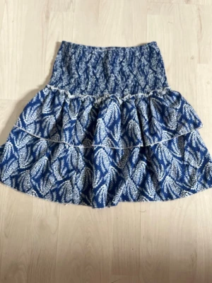 Blå volangkjol  - Kort volangkjol i blått med vitt bladmönster. Kjolen har bred smock i midjan för skön passform och dubbla volanger som ger en luftig och trendig look. Perfekt för dig som gillar en avslappnad men ändå snygg stil.