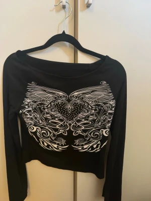 Svart långärmad topp med vingar - Svart croppad långärmad topp med vitt tryck av vingar och hjärta framtill. Toppen har en bred halsringning och är figurnära i modellen. Perfekt för dig som gillar edgy och unika detaljer.