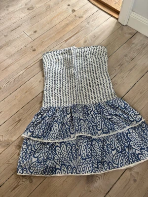 Vit och blå axelbandslös kortklänning - Snygg axelbandslös kortklänning i vitt och blått med smockad överdel och volangkjol. Klänningen har ett unikt mönster och är perfekt för varma dagar. Materialet känns lätt och luftigt, och volangerna ger en trendig look. Skriv för fler bilder 😊