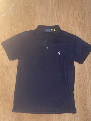 Marinblå pikétröja från Polo Ralph Lauren - Klassisk marinblå pikétröja från Polo Ralph Lauren med vit broderad logga på bröstet. Tröjan har korta ärmar, krage och knappar framtill. Perfekt till jeans eller shorts för en clean och stilren look.