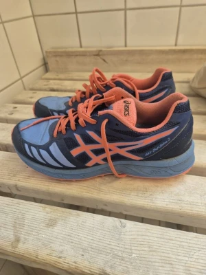 Blå och orange löparskor från Asics - Säljer ett par snygga löparskor från Asics i blått och orange. Skorna har ventilerande mesh-tyg, klassisk snörning och dämpad sula för extra komfort. Perfekta för träning eller promenader. Modellen heter Gel-Hall-Sprint 3.