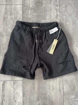 Svarta shorts från Essentials Fear of God/ Nypris 75€ - Säljer ett par svarta shorts från Essentials Fear of God med snörning i midjan och diskret logga på benet. Shortsen har en relaxed passform och är tillverkade i mjuk bomull, perfekta för en chill och stilren look. Nypris 75€
