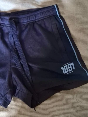 300kr! Snabb affär  - Säljer ett par marinblå shorts med snörning i midjan och vit detalj med siffran 1891 på ena benet. Shortsen har en sportig look med ljusblå rand längs sidorna och är perfekta för träning eller chill.