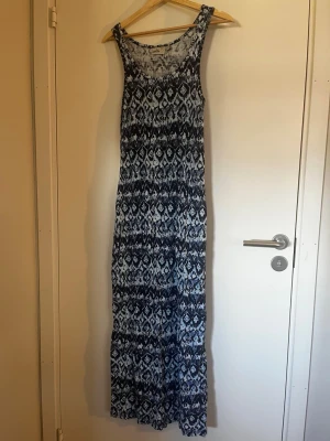 Mönstrad långklänning i blått och vitt - Långklänning med ärmlös design och rund halsringning. Klänningen har ett blått och vitt mönster som ger en bohemisk känsla. Tillverkad i mjukt och stretchigt material, perfekt för varma dagar.