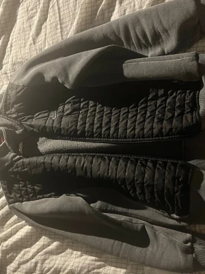 Nike cardigan - Snygg bomberjacka i svart och grått med quiltad front och släta ärmar. Jackan har ribbade muddar vid ärmslut och nederkant samt dragkedja framtill. Perfekt för dig som vill ha en stilren och modern look.