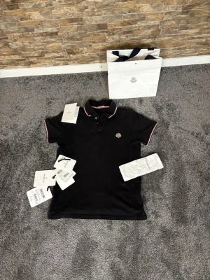 Svart Moncler pikétröja med röd/vit kant - Snygg svart pikétröja från Moncler med klassisk krage och knappar framtill. Pikén köptes för ett bra tag sen i nyskick men har till mestadels legat i en låda sen dess. Perfekt för en stilren och sportig look.