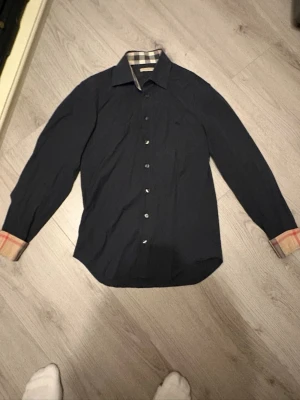 Svart skjorta från Burberry - Stilren svart skjorta från Burberry med klassiskt rutigt mönster på insidan av kragen och manschetterna. Skjortan har knappar framtill, lång ärm och diskret broderad logga på bröstet. Perfekt för dig som gillar tidlös design.