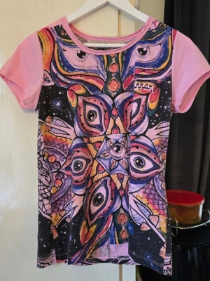 Mirror  rosa t-shirt med ögonmönster - Unik t-shirt från Mishka med psykedeliskt ögonmönster i flera färger på framsidan och svartvitt på baksidan. T-shirten har rosa korta ärmar och rund hals. Materialet känns som bomull och trycket är riktigt iögonfallande och konstnärligt.storlek M