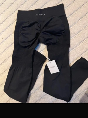 Svarta seamless leggings från Thrivin - Säljer ett par svarta seamless leggings från Thrivin med hög midja och scrunch-detalj bak. Tillverkade i stretchigt material som formar sig efter kroppen. Perfekta för träning eller chill. Diskret logga i midjan och ribbade partier för extra komfort.