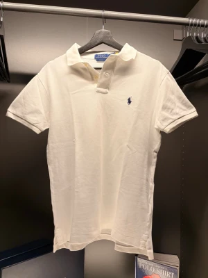  Ralph Lauren - Klassisk vit pikétröja från Ralph Lauren i slim fit-modell. Tröjan har korta ärmar, krage och knappar framtill samt den ikoniska broderade loggan på bröstet. Tillverkad i mjuk bomull som känns skön mot huden.