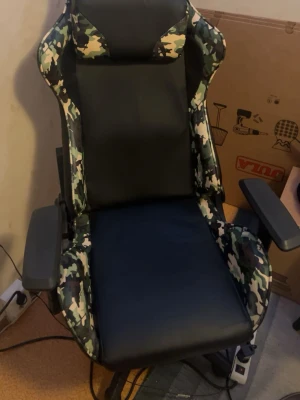 Gamingstol svart och camo - Säljer en gamingstol med svart sits och ryggstöd i konstläder samt camouflagemönstrade detaljer på sidorna. Stolen har justerbara armstöd och hög rygg för extra komfort. Perfekt för långa spelsessioner eller som kontorsstol.