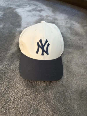New York Yankees keps från '47 Brand - Snygg New York Yankees keps från '47 Brand med vit krona och mörkblå skärm. Klassisk böjd skärm och broderad NY-logga framtill i mörkblått. Tillverkad i bomullstyg och har ventilerande hål upptill. Perfekt för en sportig och avslappnad stil.