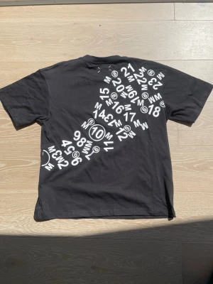 MM6 Maison Margiela x Tommy Cash Collab Tee – Black – Size S – Brand New with tags - MM6 Maison Margiela x Tommy Cash collaboration t-shirt in black. Overlapping logos on the chest, scattered numbers graphic on the back. Brand new with tags, never worn. Size S but fits oversized – works well on M too. Rare collab piece. Selling for 500 SEK. Shipping not included, local pickup possible.
